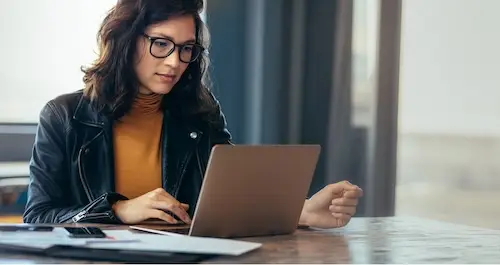 Woman with glasses, medium length brown hair, looking at her laptop.