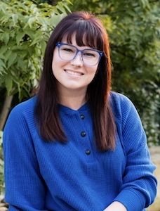 Headshot photo of clinician Corinne sitting outside in front of a tree. She is smiling, wearing a blue shirt, has blue glasses, and long straight brown hair.