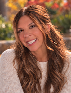 woman with long wavy brown hair, smiling with head tilted left; wearing light pink sweater, professional portrait