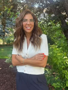 Female therapist headshot outdoors with greenery in the background. She is wearing a white blouse, glasses, has long brown wavy hair, and has arms crossed.