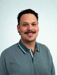 Male counselor headshot with light blue background. Subject is smiling, has short brown curly hair, mustache, and a green polo shirt.