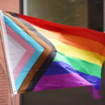 Progress pride flag flowing in the wind with a pink building and dark window in the background.