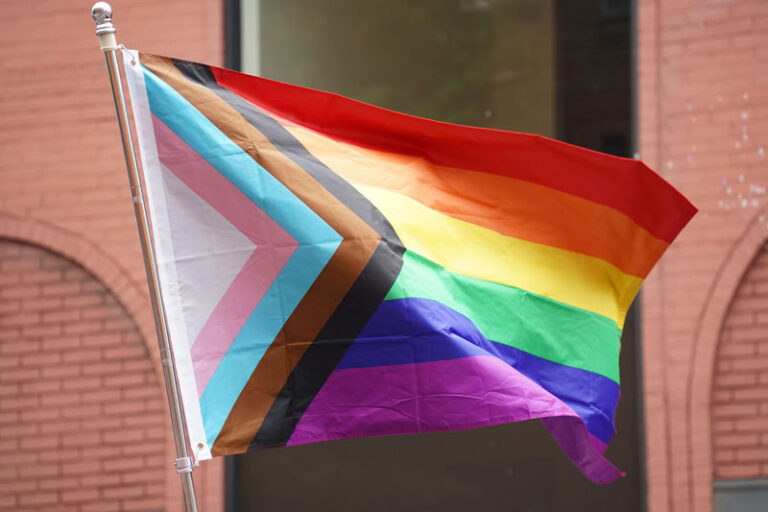 Progress pride flag flowing in the wind with a pink building and dark window in the background.
