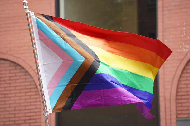 Progress pride flag flowing in the wind with a pink building and dark window in the background.