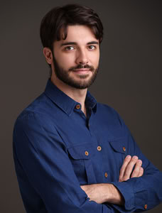 Male counselor headshot with grey background. Subject is wearing a navy blue button up shirt with arms crossed. He has a neutral look and has a full beard with short brown hair.
