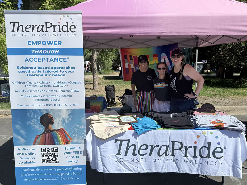 TheraPride Boulder, CO pride 2026 booth. Vertical sign on the left advertising services. Pink colored tent in the center and right top of the screen with a white table and cloth at the bottom, and 3 female therapists in the middle smiling.